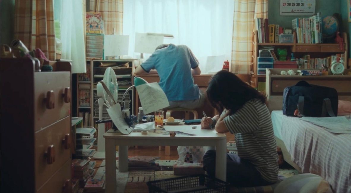 Fujino sits at her desk while Kyomoto works at the table on the ground